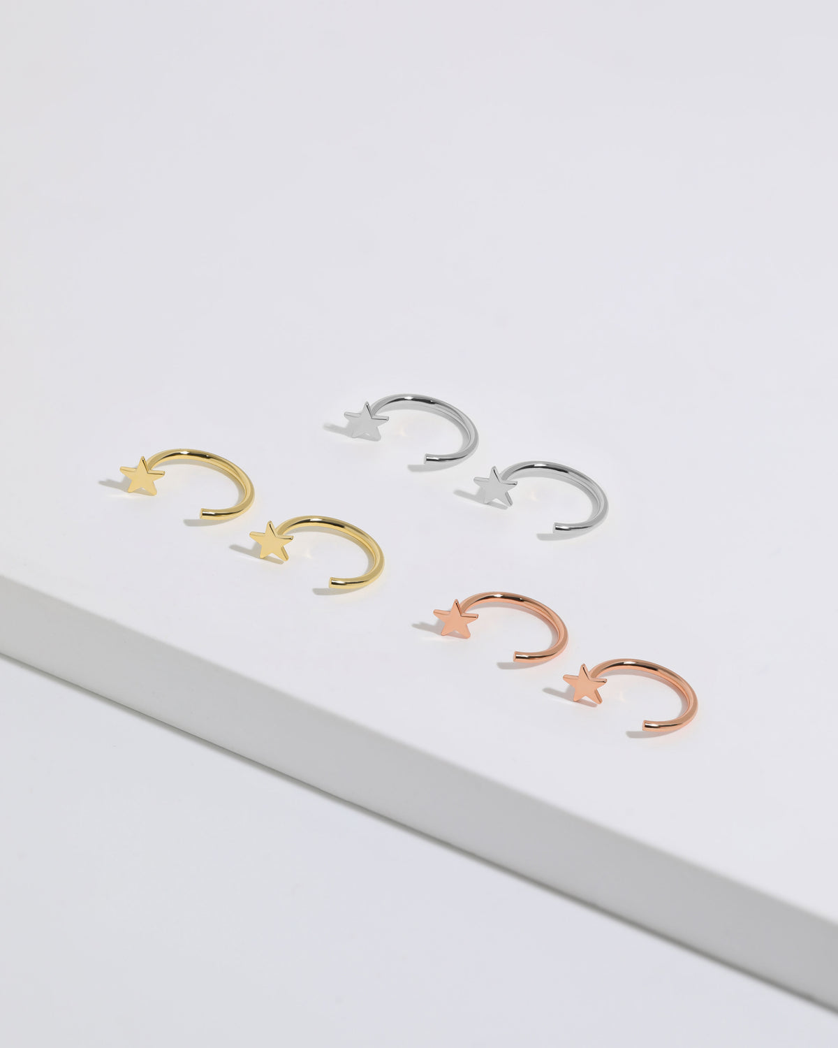 Star Arc Huggie Hoop Earrings in silver, gold, and rose gold colors arranged together, featuring polished stars and open curved hoops.