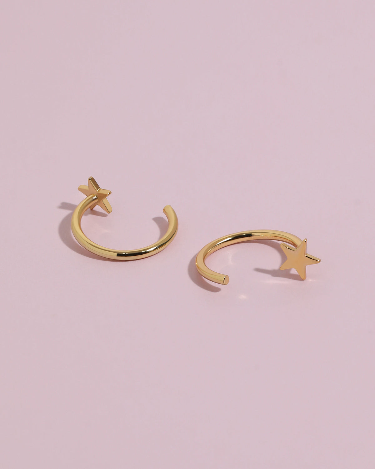 Star Arc Huggie Hoop Earrings in gold color resting on a soft pink background, showing the polished star detail and open hoop construction.