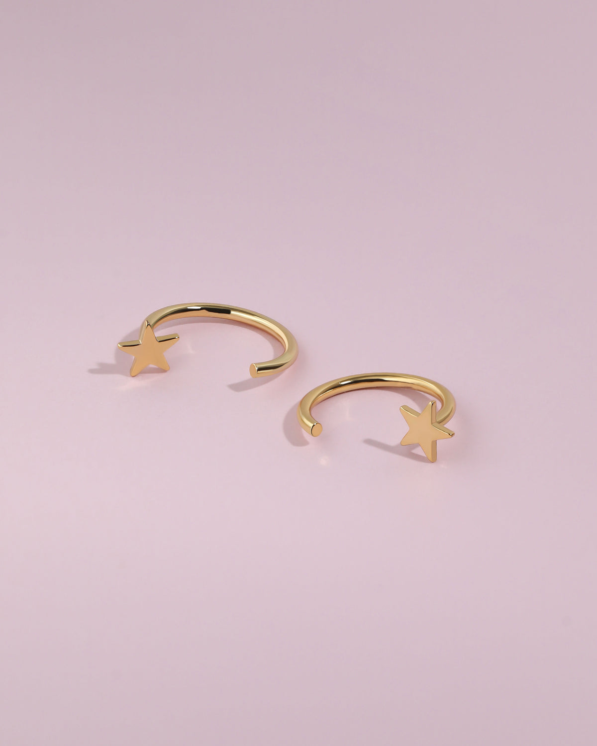 Pair of Star Arc Huggie Hoop Earrings in gold color displayed flat on a pink background with polished star details.
