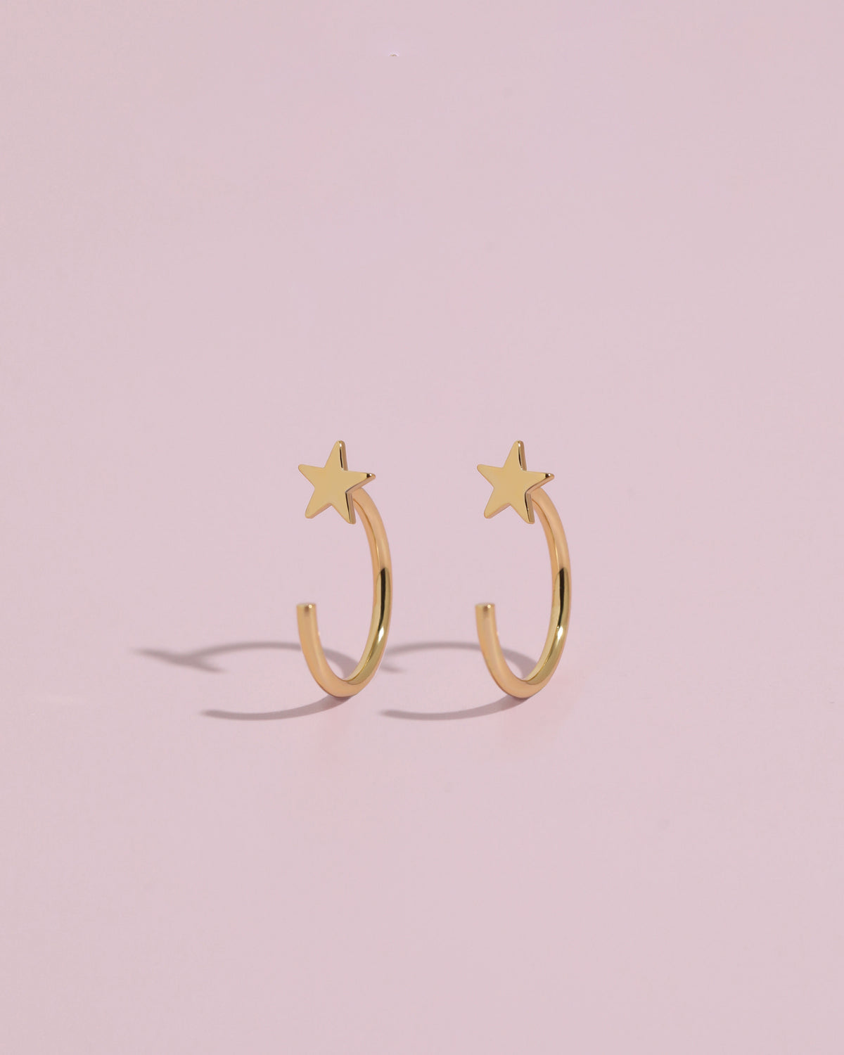 Pair of Star Arc Huggie Hoop Earrings in gold color standing upright on a pink background, highlighting the curved hoop and star design.