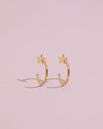 Pair of Star Arc Huggie Hoop Earrings in gold color standing upright on a pink background, highlighting the curved hoop and star design.