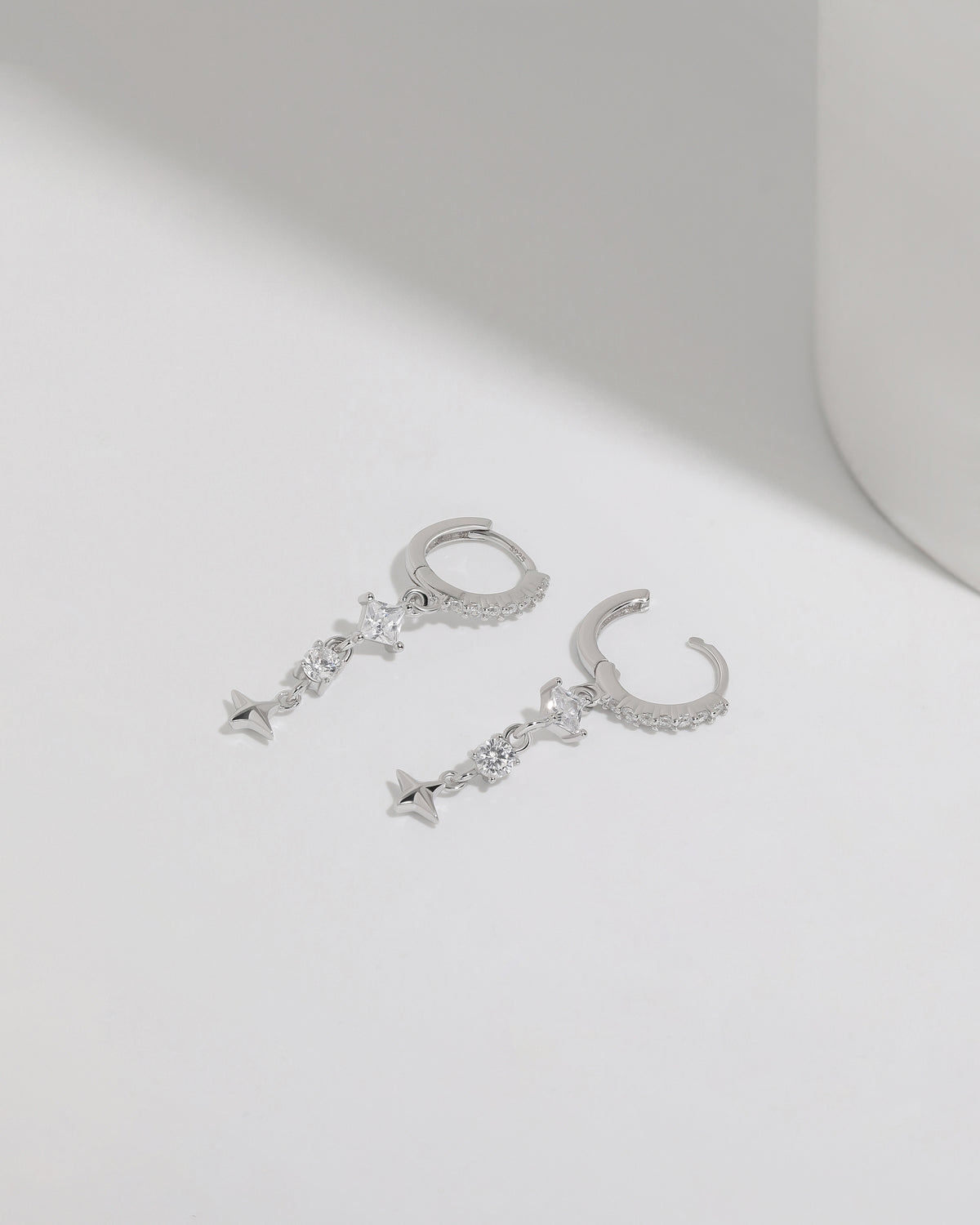 Starlight Drop Huggie Earrings shown as a pair on a white surface, with the hoops open to highlight the clasp and dangling star charm design.