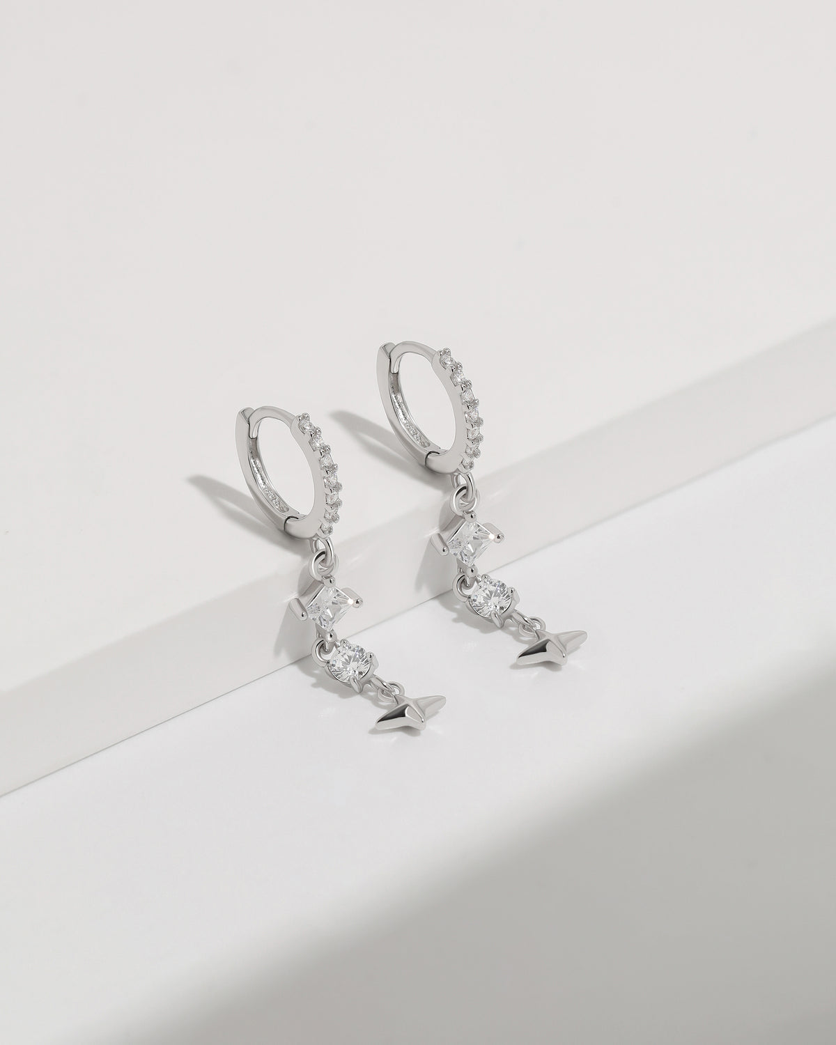Starlight Drop Huggie Earrings displayed hanging as a pair, featuring pavé hoops, geometric drop stones, and a polished star charm.