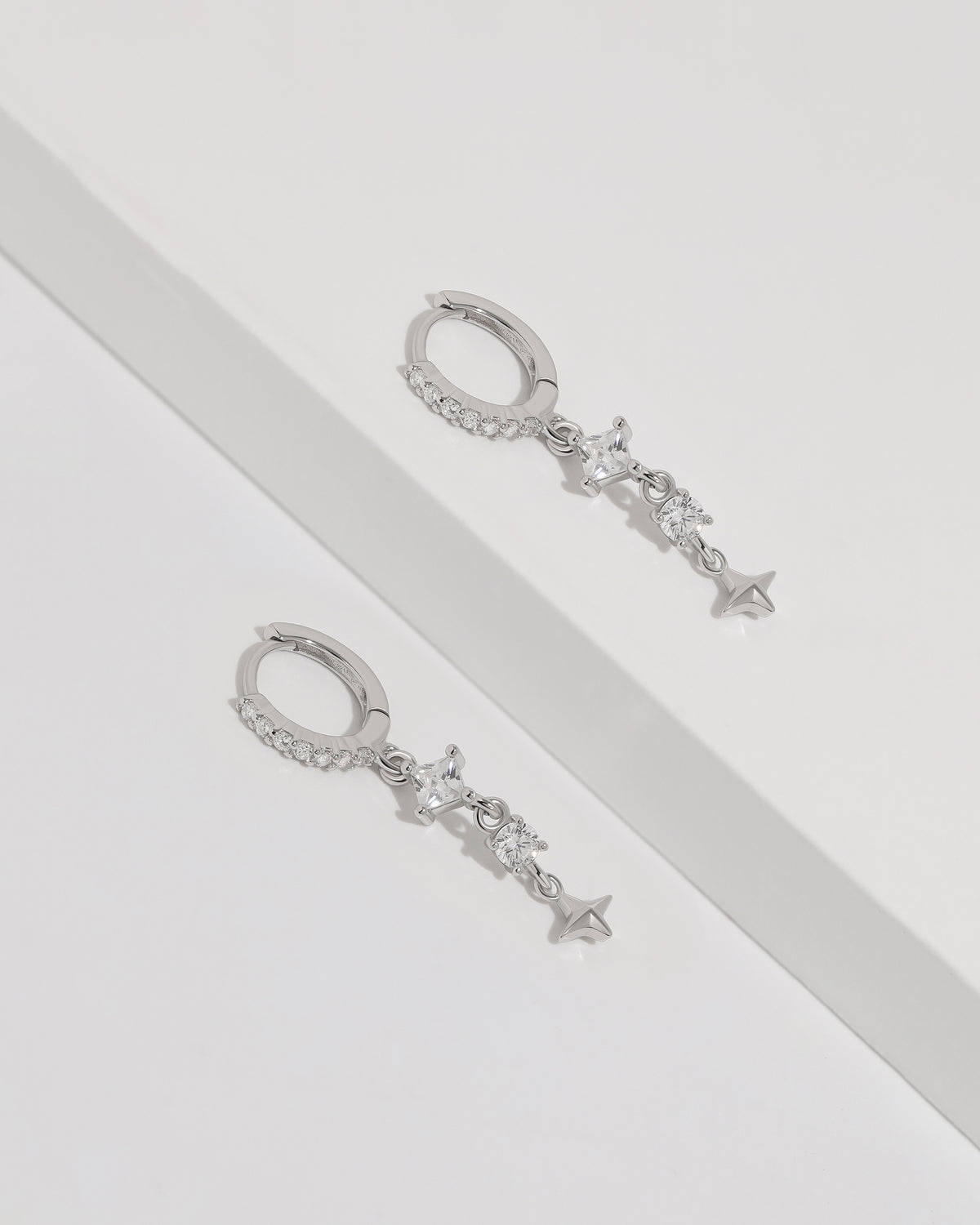 Starlight Drop Huggie Earrings laid flat as a pair on a white surface, showing the slim huggie hoops and delicate star drop details.