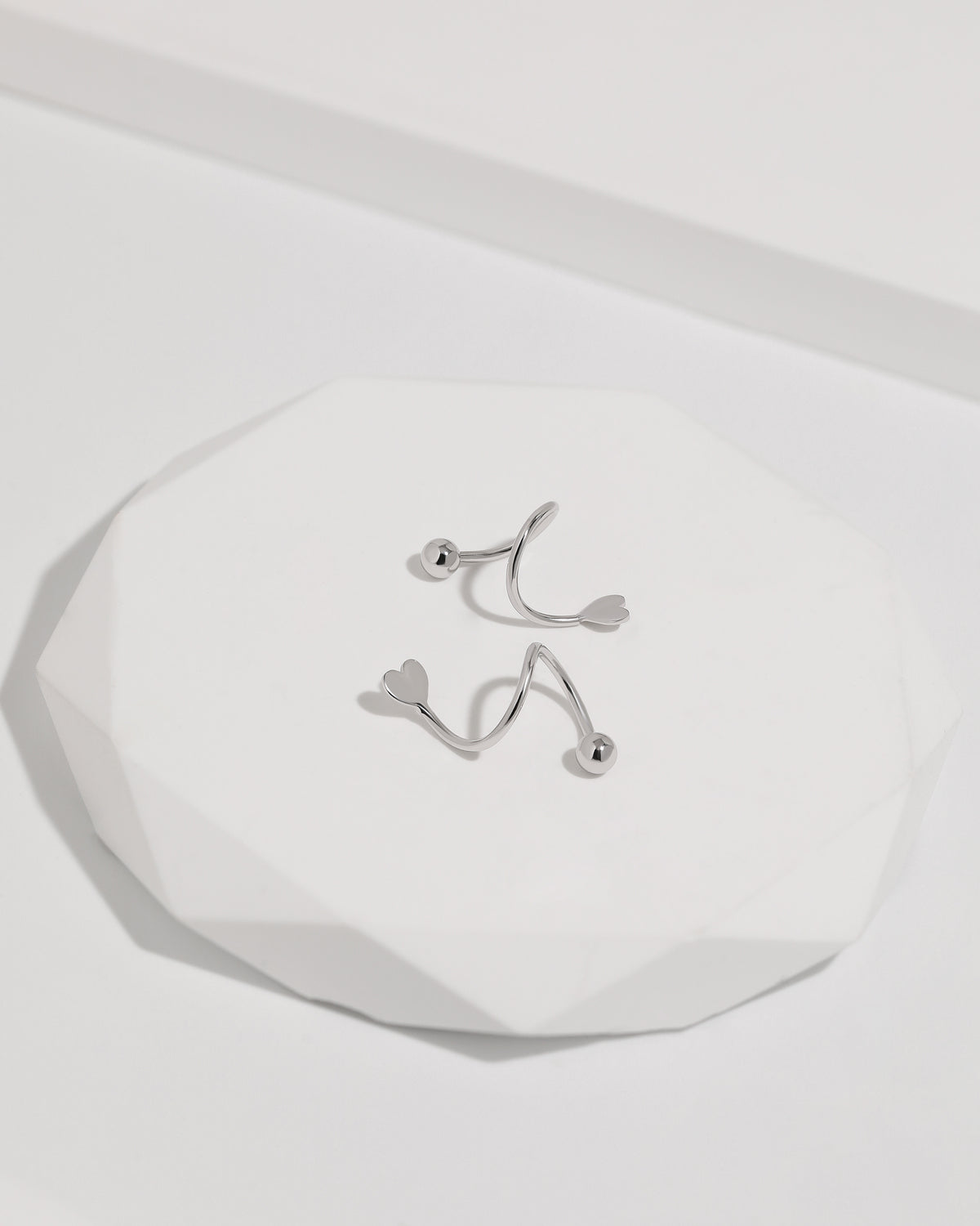 Heart Spiral Screw Back Earrings in silver color displayed on a white geometric surface, showing the sculptural spiral shape, heart ends, and polished round screw back balls.