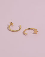 Star Arc Huggie Hoop Earrings in gold color resting on a soft pink background, showing the polished star detail and open hoop construction.