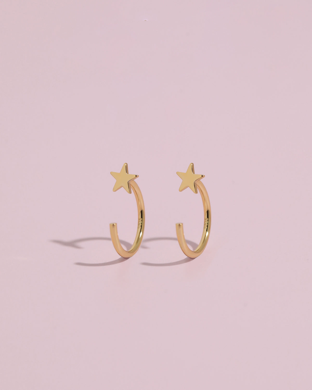 Pair of Star Arc Huggie Hoop Earrings in gold color standing upright on a pink background, highlighting the curved hoop and star design.