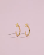 Pair of Star Arc Huggie Hoop Earrings in gold color standing upright on a pink background, highlighting the curved hoop and star design.