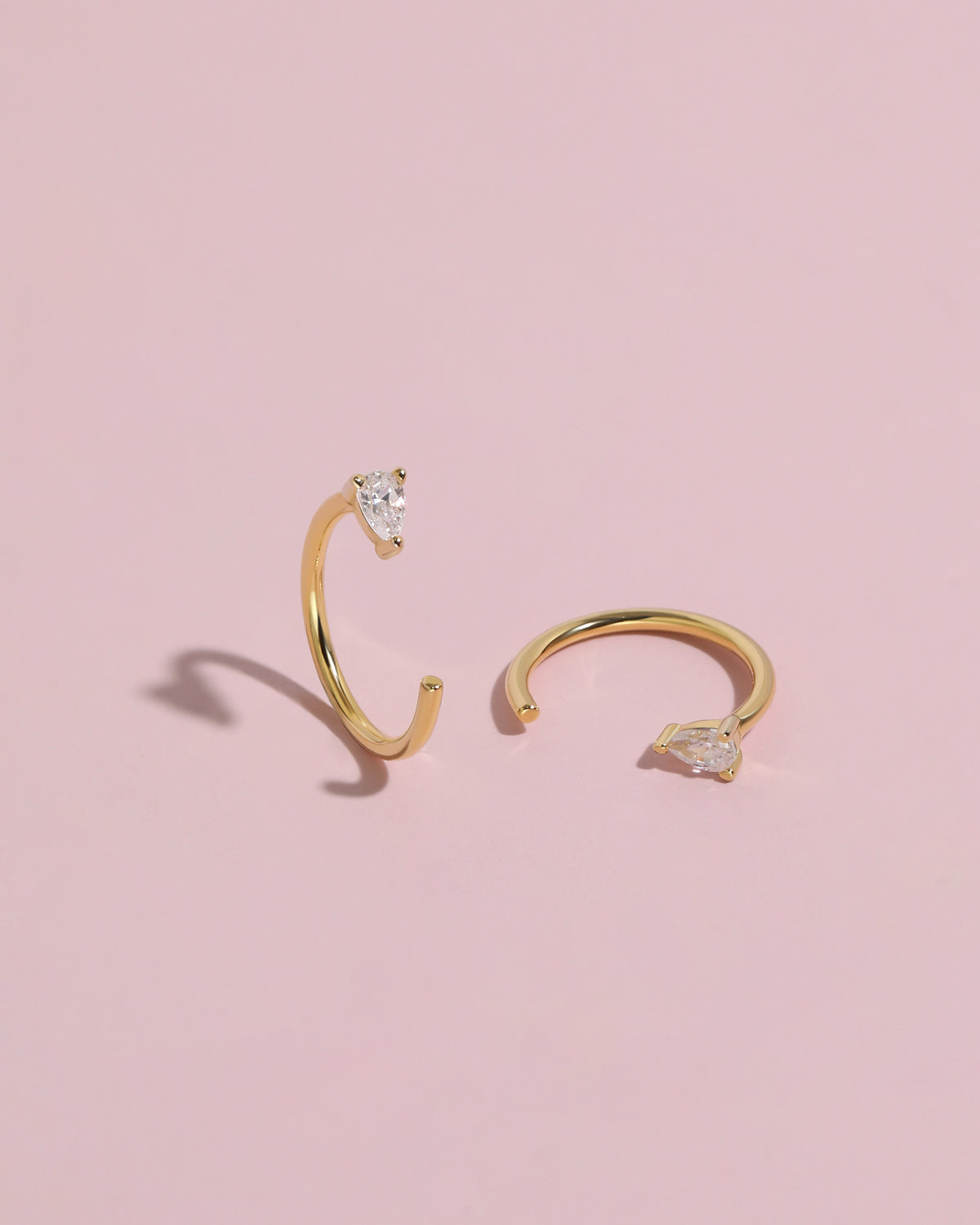Pair of gold-tone Meissa Pear Sparkle Huggie Hoop Earrings with clear pear-cut stones on a soft pink background.