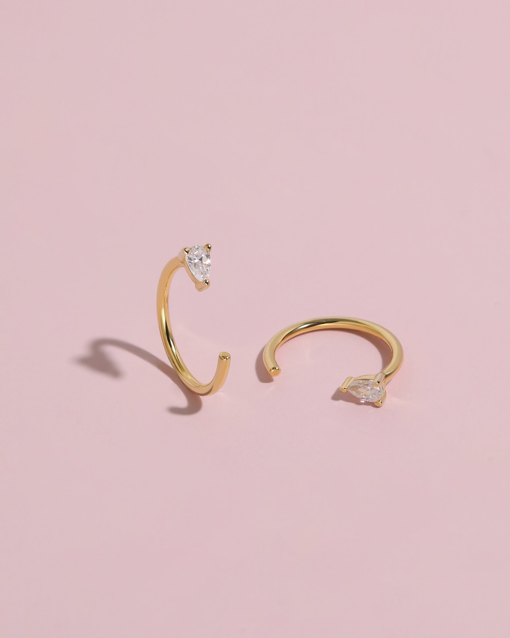 Pair of gold-tone Meissa Pear Sparkle Huggie Hoop Earrings with clear pear-cut stones on a soft pink background.