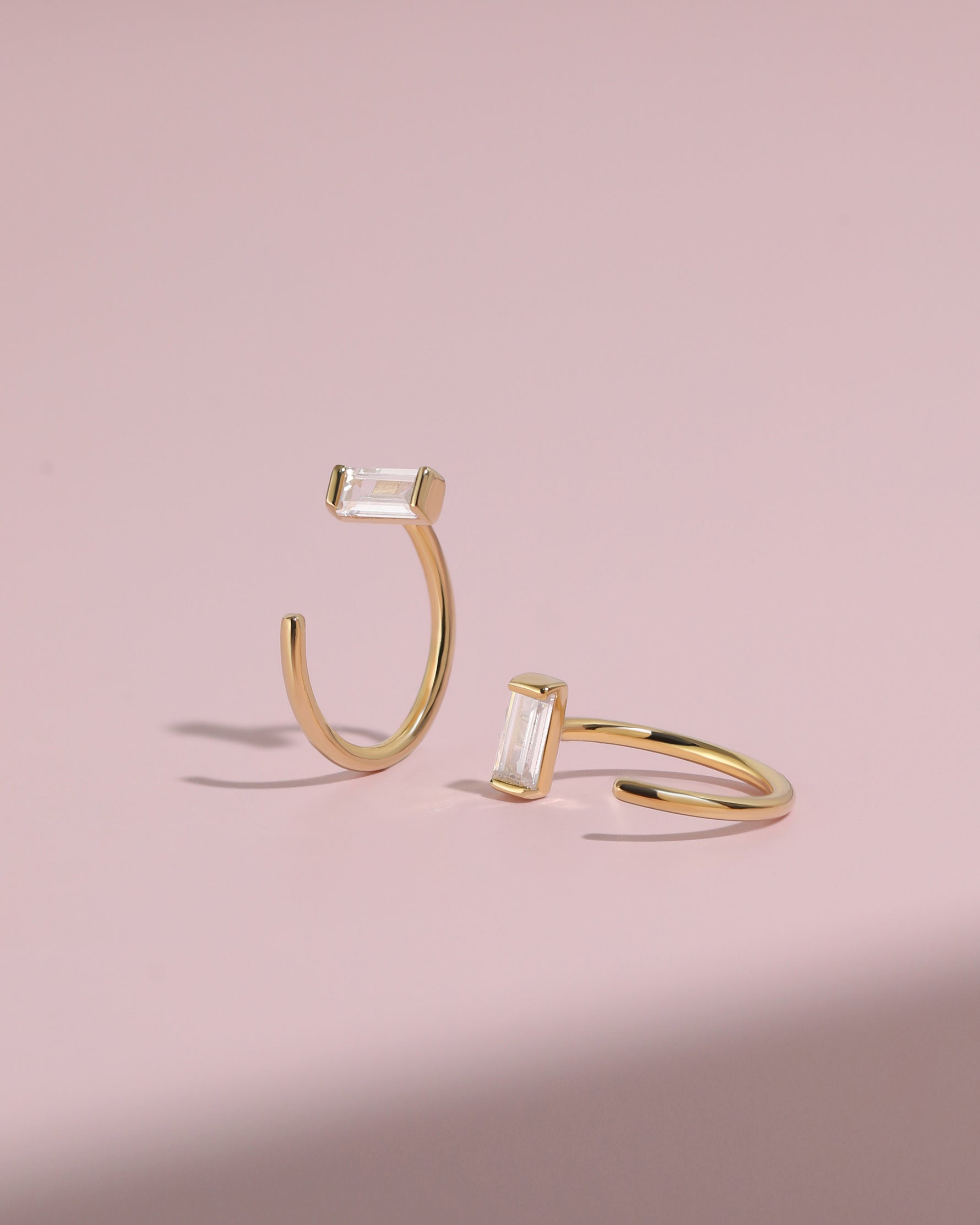 Pair of gold-tone Meissa Baguette Sparkle Huggie Hoop Earrings with clear baguette-cut stones on a soft pink background.