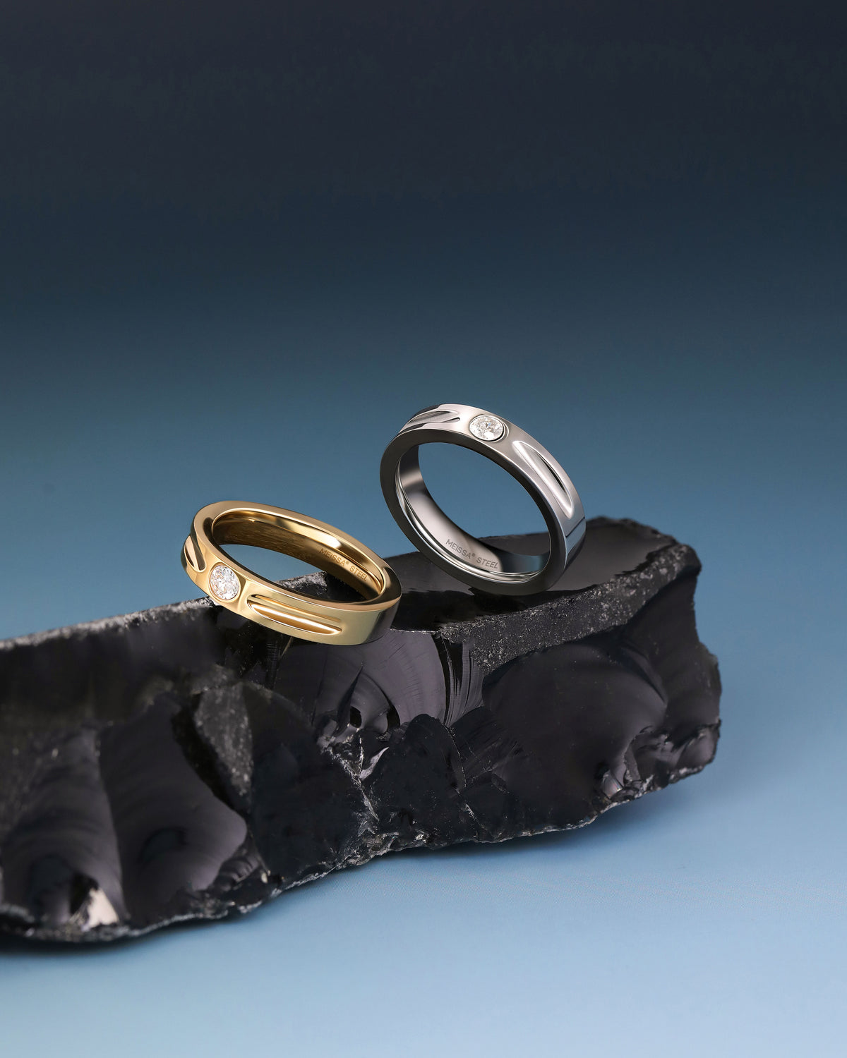 Gold and silver Nova Track Rings in stainless steel displayed on black stone against a blue background.