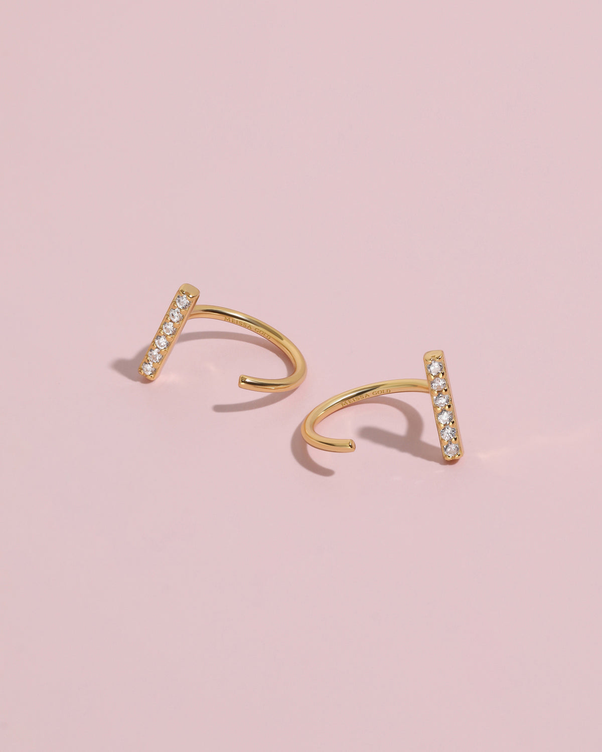 Pair of gold-tone Meissa Sparkling Bar Huggie Hoop Earrings with clear stones on a soft pink background.