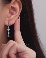Starlight Drop Huggie Earrings shown worn on the ear and held by hand, featuring sparkling pavé huggie hoops with square and round stones, delicate dangling links, and polished star charms, with close-up views highlighting the drop length, movement, and size.